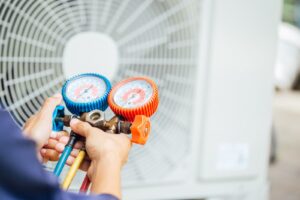 Two hands holding a manifold gauge set with a blurry outdoor HVAC unit in the background.