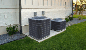Two AC units outside of a white house, one twice as tall as the other, and a green lawn next to them.